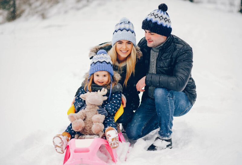Family have fun in a winter park. Stylish mother in a blue jacket. Little girl on a pink sled. Father with cute daughter